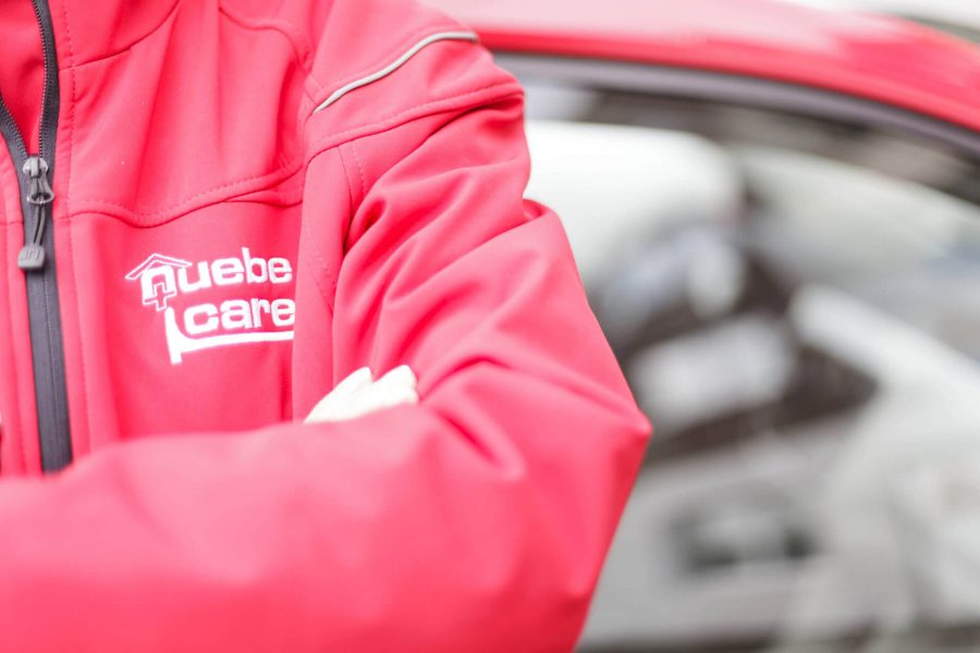 Nahaufnahme einer Person in roter Jacke mit dem Logo „Quebe Care“. Im Hintergrund ist ein rotes Auto mit geöffneter Tür zu sehen.